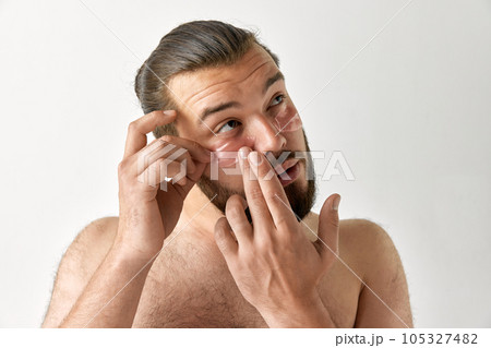 Man looking up and puts patch on area under eyes over isolated grey background. Bearded man. Man looking up and puts patch on area under eyes over isolated grey background. Bearded man. 105327482
