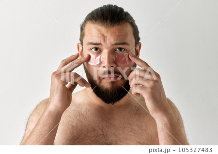 Photoshoot of man looking at camera grimacing and applying eye patches over isolated grey background. Photoshoot of man looking at camera grimacing and applying eye patches over isolated grey background. 105327483