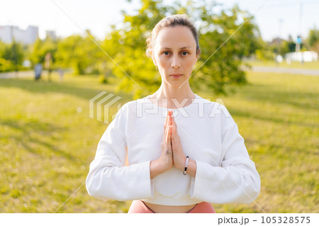 Portrait of peaceful young woman wearing sportswear standing in namaste position and looking at camera relaxing at city park on sunny summer day. Concept of practicing yoga alone outdoors. Portrait of peaceful young woman wearing sportswear standing in namaste position and looking at camera relaxing at city park on sunny summer day. Concept of practicing yoga alone outdoors. 105328575