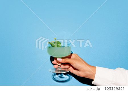 Female hand holding glass with cocktail on bright pop art blue background with shadow. Copy space for ad. Female hand holding glass with cocktail on bright pop art blue background with shadow. Copy space for ad. 105329893