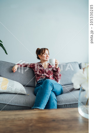 Young smiling woman sitting on sofa and looking side up while drinking coffee. Young brunette woman relaxing after housekeeping, home cleaning. Portrait of relaxed female resting at home. Vertical. 105330285