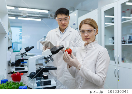 Portrait of an Asian man and a female nurse working together in a laboratory with microscopes. They eat food, hold vegetables in their hands. They look seriously into the camera. 105330345