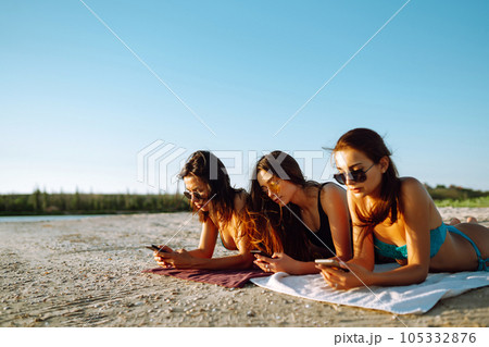 Three beautiful girls using mobile smart phone at the beach. Girls sunbathing on the beach. Three beautiful girls using mobile smart phone at the beach. Girls sunbathing on the beach. 105332876