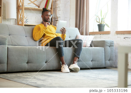Young african an, freelancer sitting on sofa at home and having online business meeting via ablett, meeting with colleagues 105333196