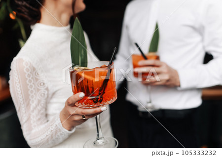 hands of woman and man are clinking, cheers with glasses of Spritz cocktail. Couple celebrating wedding, anniversary with Aperol spritz cocktails, with orange and greens Refreshing alcoholic drink 105333232