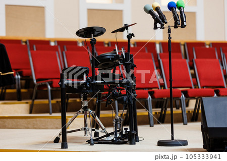 Coloured microphones on stand and electronic drum set on podium, a lot of red chairs blurred in the background. Concept of concert, event, communication, seminar, media, graduation, performance 105333941