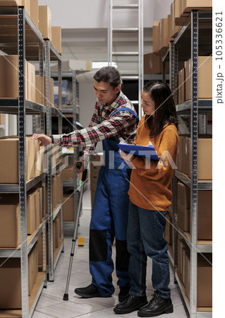 Delivery manager coordinating warehouse worker picking order and searching parcel in storage. Storehouse employee on crutches checking product in stock and cooperating with distribution operator Delivery manager coordinating warehouse worker picking order and searching parcel in storage. Storehouse employee on crutches checking product in stock and cooperating with distribution operator 105336621