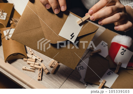 Unrecognizable young woman sticks number on craft bag, fastens with clothespin. Female making kraft paper for homemade advent calendar Made with your own hands step by step DIY crafts do it yourself 105339103