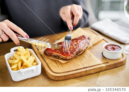Lunch in a restaurant, a woman cuts Delicious Pork ribs. Full rack of ribs BBQ on wooden plate with french fries and salad Lunch in a restaurant, a woman cuts Delicious Pork ribs. Full rack of ribs BBQ on wooden plate with french fries and salad 105348570