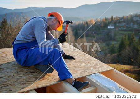 Carpenter hammering nail into OSB panel on the roof top of future cottage. Man worker building wooden frame house. Carpentry and construction concept. Carpenter hammering nail into OSB panel on the roof top of future cottage. Man worker building wooden frame house. Carpentry and construction concept. 105351873