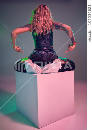 Young fitness blonde woman wearing blue velour booty shorts posing on camera, sits on top of big cube, back view Young fitness blonde woman wearing blue velour booty shorts posing on camera, sits on top of big cube, back view 105352621