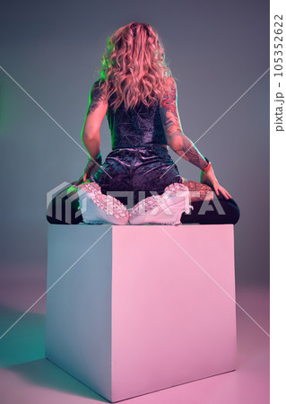 Young fitness blonde woman wearing blue velour booty shorts posing on camera, sits on top of big cube, back view 105352622