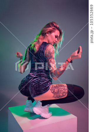 Young fitness blonde woman wearing blue velour booty shorts posing on camera, making squats on top of big cube 105352669