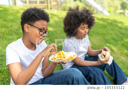 Dark skinned friends or sister and brother sitting on grass in park and eating junk food enjoying. Dark skinned friends or sister and brother sitting on grass in park and eating junk food enjoying. 105353122