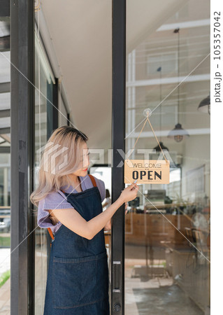 Young asian businesswoman small business owner standing at door entrance signboard, A cheerful entrepreneur young waitress in a blue apron near the glass door 105357042