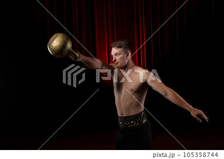 Power gymnast circus with weight.Handsome guy with a muscular body holding a kettlebell on a black stage background 105358744