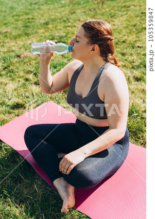 Attractive young 30s girl finished her work out and now drinking water in nature. She is sitting on a yoga mat on green grass. 105359847
