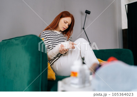 Side view of suffering from pain injured young woman with broken right hand wrapped in white gypsum bandage sitting on couch. Sad redhead female with broken arm from accident at home alone. 105364023