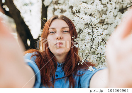 Young happy woman is making selfie on a camera on city street. Girl talking on a video call with phone outdoor. White flowers in the tree in the early spring. Flowers of the cherry blossoms 105366962