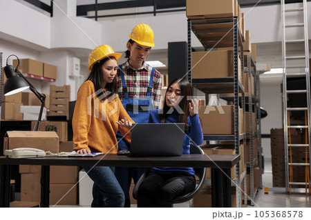 Asian distribution center workers checking goods supply schedule on laptop. Warehouse woman manager sitting at desk while explaining logistics chain to coworkers in industrial storage room 105368578