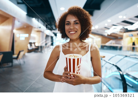 Curly-haired woman with an afro hairstyle in a white shirt is walking to watch a movie with popcorn in hand 105369321