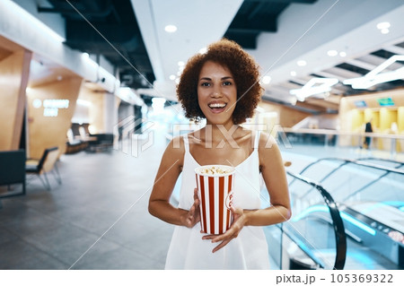 Curly-haired woman with an afro hairstyle in a white shirt is walking to watch a movie with popcorn in hand Curly-haired woman with an afro hairstyle in a white shirt is walking to watch a movie with popcorn in hand 105369322