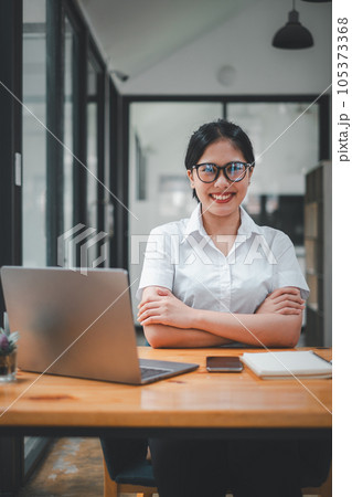 Portrait of young smiling woman looking at camera with crossed arms. Happy girl sitting in creative office. Successful businesswoman standing in office with copy space. Portrait of young smiling woman looking at camera with crossed arms. Happy girl sitting in creative office. Successful businesswoman standing in office with copy space. 105373368