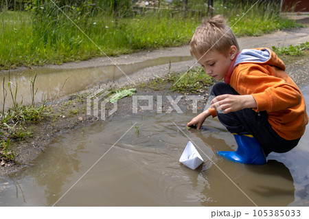 An 8-year-old boy in an orange sweatshirt lets paper boat into puddle while squatting on summer day. Games alone. Copy space 105385033