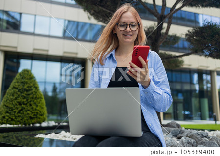 Happy Caucasian student freelancer blonde woman rest use laptop pc computer talk by mobile cell phone look, sitting outdoors against blurred city building. She is keyboarding on laptop. Freelance work Happy Caucasian student freelancer blonde woman rest use laptop pc computer talk by mobile cell phone look, sitting outdoors against blurred city building. She is keyboarding on laptop. Freelance work 105389400