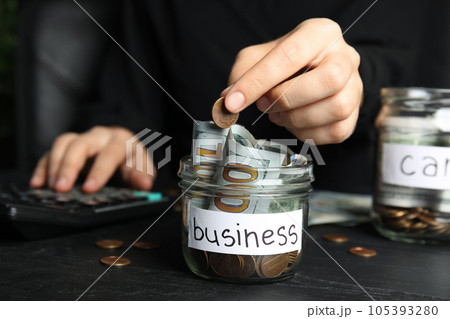 Woman putting coin into jar with money and tag BUSINESS on black table, closeup 105393280