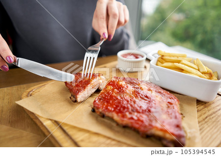 Lunch in a restaurant, a woman cuts Delicious Pork ribs. Full rack of ribs BBQ on wooden plate with french fries and salad 105394545
