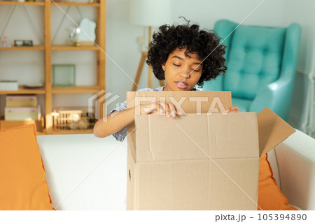 African girl unpacking delivery looking in box. Happy woman opening carton box. Female getting parcel looking at delivered goods items. Satisfied client positive feedback quick express deliver 105394890