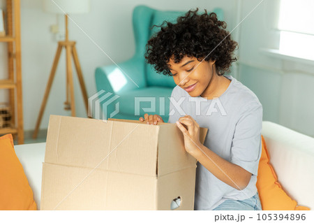 African girl unpacking delivery looking in box. Happy woman opening carton box. Female getting parcel looking at delivered goods items. Satisfied client positive feedback quick express deliver African girl unpacking delivery looking in box. Happy woman opening carton box. Female getting parcel looking at delivered goods items. Satisfied client positive feedback quick express deliver 105394896