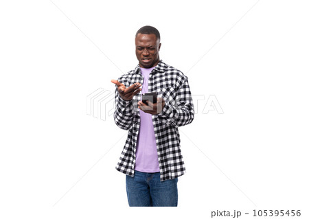 portrait of a confused dark-skinned man in a plaid shirt and jeans holding a smartphone in his hand 105395456