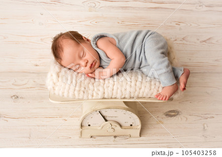 A cute newborn baby boy sleeps in a blue overalls in the first days of life. Against a background of light white wood. On the scales for weighing newborns. Professional portrait photography A cute newborn baby boy sleeps in a blue overalls in the first days of life. Against a background of light white wood. On the scales for weighing newborns. Professional portrait photography 105403258