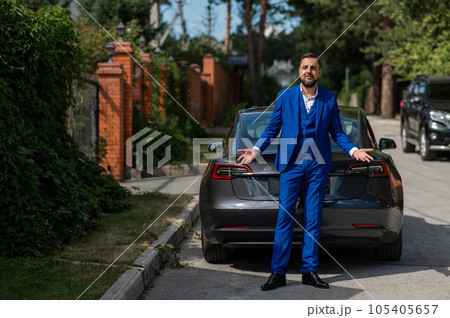 Caucasian bearded man in a blue suit stands near a black car in the countryside in summer. 105405657