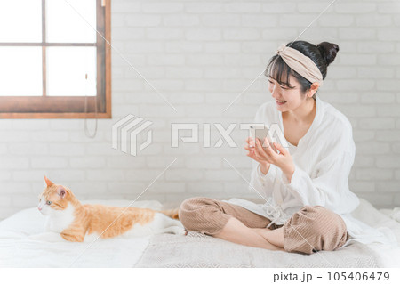 家のベッドルームでペットの猫とスマホで写真を撮って遊ぶアジア人女性 105406479