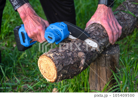 Hand-held portable camping electric saw on a battery for cutting firewood and wood. Close-up, a hand sawing a log, splinters flying Hand-held portable camping electric saw on a battery for cutting firewood and wood. Close-up, a hand sawing a log, splinters flying 105409500