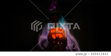 Witch with blue and violet hair, black hat with violet ribbon and long black nails is holding pumpkin in her hands. 105411912