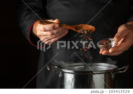The chef adds fragrant dry peppers to a pot of boiling food. Restaurant kitchen cooking concept with advertising space on black background. Spoon in the cook hand. The chef adds fragrant dry peppers to a pot of boiling food. Restaurant kitchen cooking concept with advertising space on black background. Spoon in the cook hand. 105413529