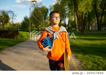 Portrait of little boy carrying helmet and skateboard over street background Portrait of little boy carrying helmet and skateboard over street background 105417689