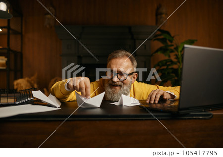 Happy excited mature grey-haired man playing with paper boat at workplace Happy excited mature grey-haired man playing with paper boat at workplace 105417795