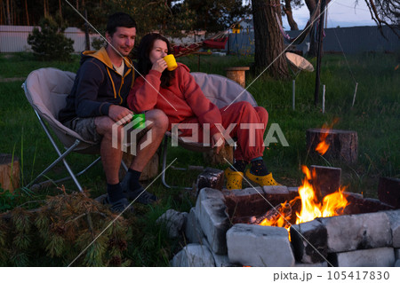Couple in love is sitting by the outdoor campfire in the courtyard of the house on camping chairs, a romantic evening. a man and a woman warm their hands by the fire, drink tea 105417830