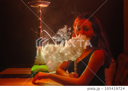 A sexy young woman smokes a hookah and releases a cloud of steam through her nostrils. Close up. Red tint. Night lifestyle 105419724