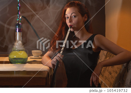 Young caucasian woman smokes a hookah or shisha in the night club or bar smoke. Smoke and steam in the air. Close up 105419732