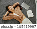Young female in casual wear smiling with closed eyes while lying on comfortable bed with opened laptop and notebook near her. 105419997