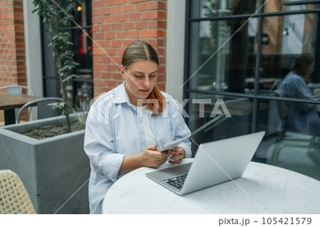 Caucasian student freelancer woman rest use laptop pc computer talk by mobile cell phone look, sitting at city cafe. She is keyboarding on laptop. Freelance work concept. Caucasian student freelancer woman rest use laptop pc computer talk by mobile cell phone look, sitting at city cafe. She is keyboarding on laptop. Freelance work concept. 105421579