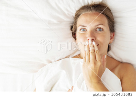 Young woman lies in bed and covers her mouth with her hand Young woman lies in bed and covers her mouth with her hand 105431692