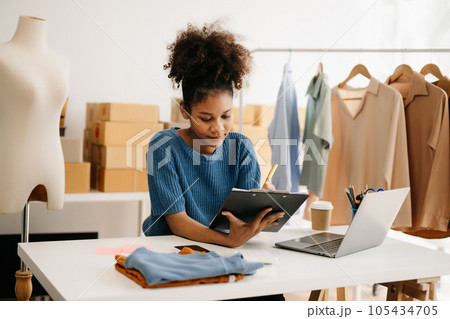 Calm curly brunette dark skinned woman on desk in modern office of fashion designer and holds tablet and smartphone. 105434705
