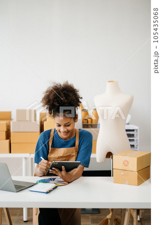 Startup small business SME, Entrepreneur owner African woman using smartphone or tablet taking receive and checking online purchase shopping order to preparing pack product box. in office. Startup small business SME, Entrepreneur owner African woman using smartphone or tablet taking receive and checking online purchase shopping order to preparing pack product box. in office. 105435068